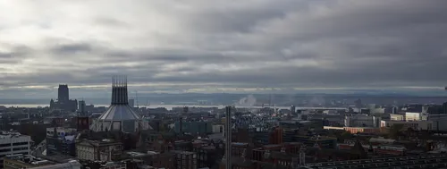 This urban landscape photograph captures a wide view of Liverpool in the United Kingdom during a late autumn morning. The main subjects are the prominent churches: the Liverpool Metropolitan Cathedral with its distinctive modern architecture, and the traditional Liverpool Cathedral, both visible against the city skyline. Low clouds cover the sky, with hints of morning light breaking through, and smoke or mist can be seen rising in the distance. The River Mersey and the distant Welsh hills are visible in the background, adding depth to the scene. The image showcases the blend of historic and contemporary architecture that defines Liverpool, highlighting both the Liverpool Cathedral and Liverpool Metropolitan Cathedral as significant landmarks within this urban setting.